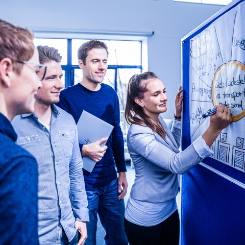 Équipe en atelier devant un tableau blanc, une collaboratrice prend des notes